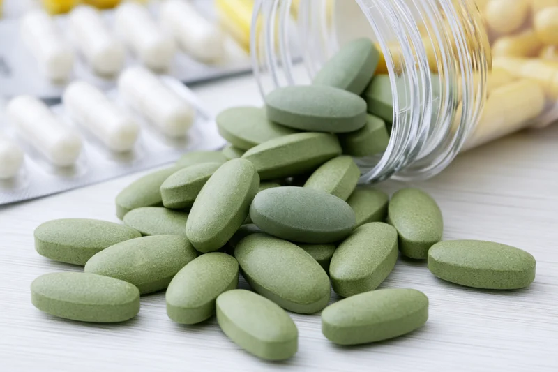 Green tablets spilling from a clear plastic bottle onto a white wooden surface