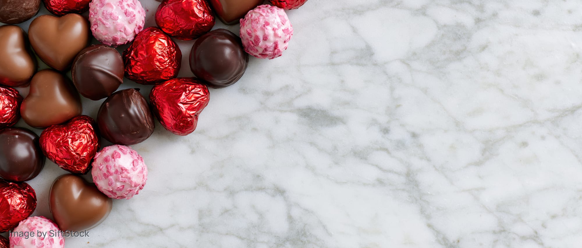 Heart-shaped and round chocolates in red, pink, and brown foil arranged along the left side of a white marble surface