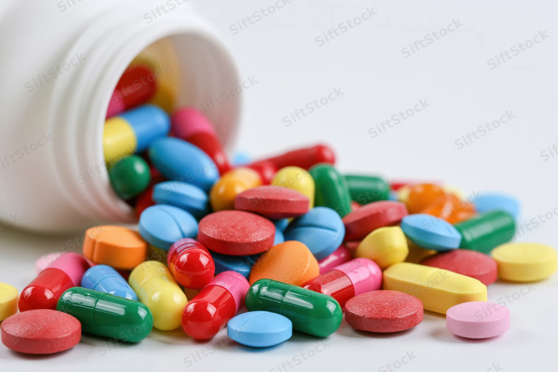 Colorful pills and capsules spilled from a white bottle on a plain surface