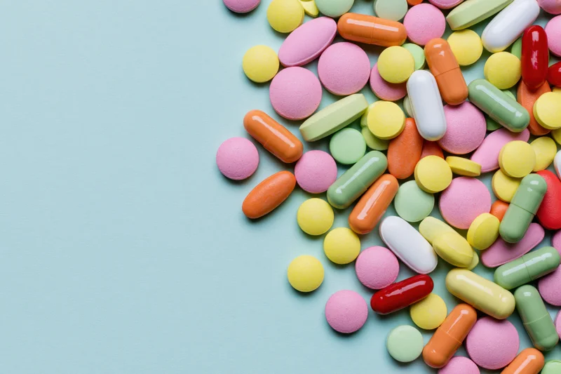 Colorful pills and capsules scattered on a light blue surface with copyspace on the left