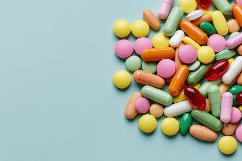 Colorful pills and capsules scattered on a light blue surface with copyspace on the left