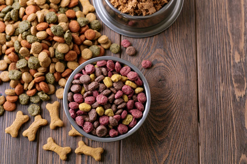 Assorted dry dog food kibble and bone-shaped treats scattered on a wooden table with metal bowls