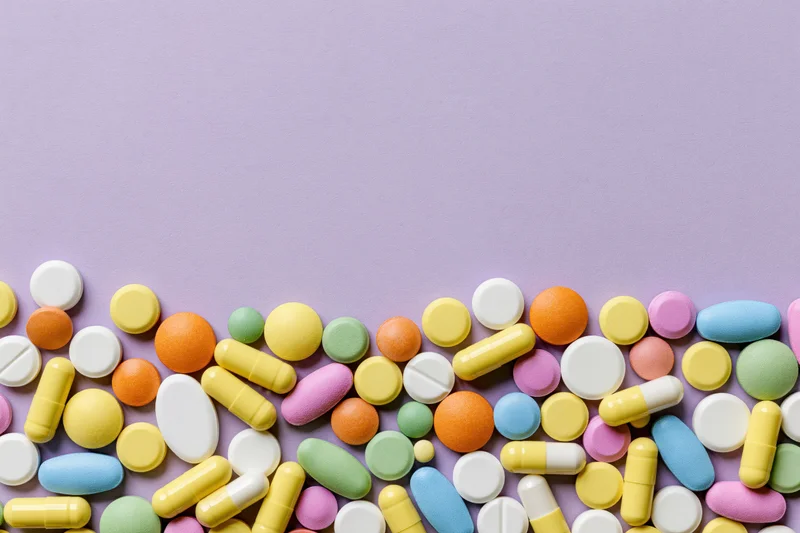 Assorted pharmaceutical pills and capsules scattered across a purple surface in a flat lay arrangement