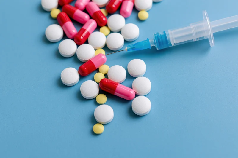 Assorted pharmaceutical pills and capsules scattered beside a syringe on a blue surface