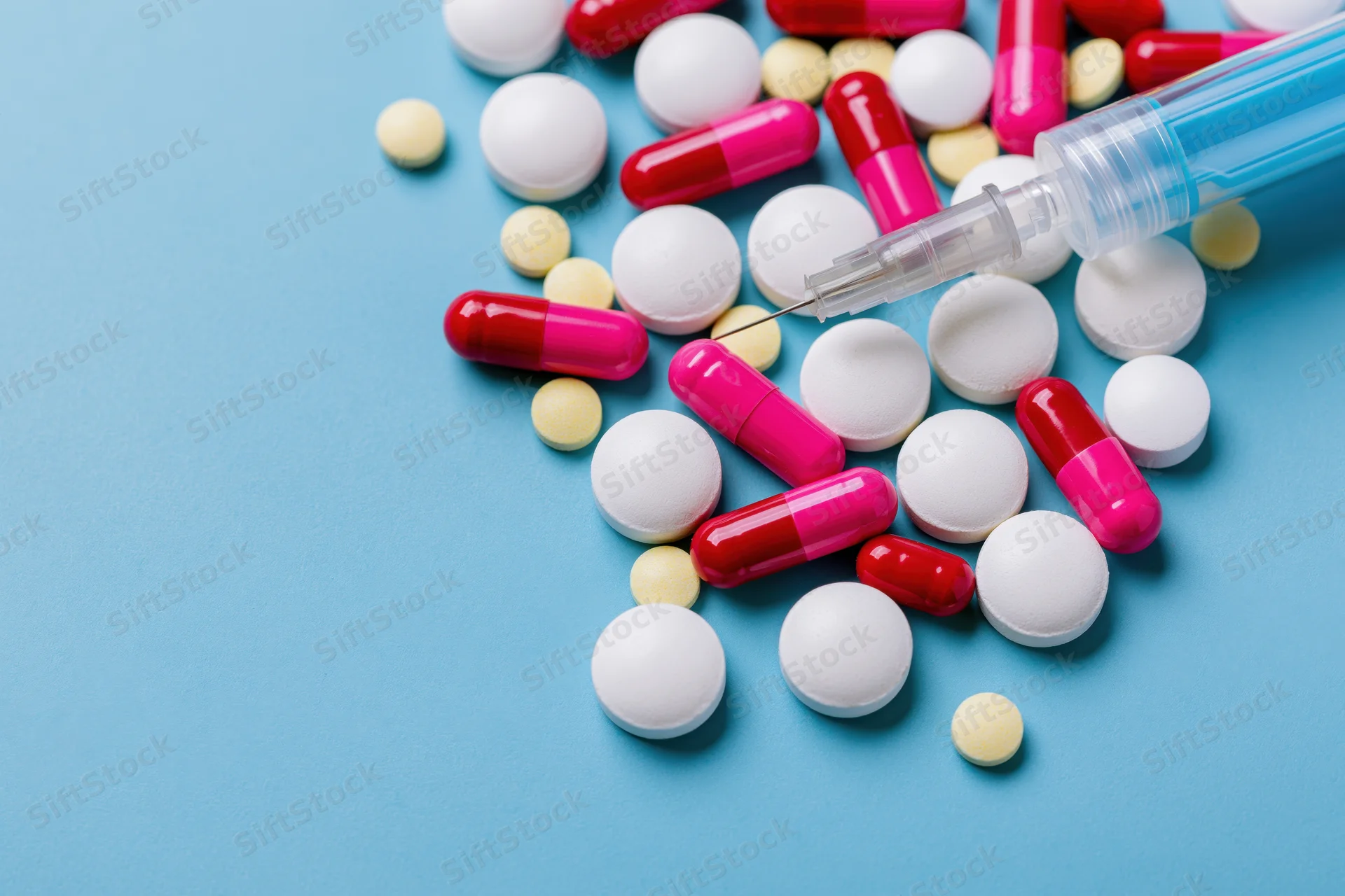 Assorted pharmaceutical pills and capsules scattered beside a syringe on a blue surface