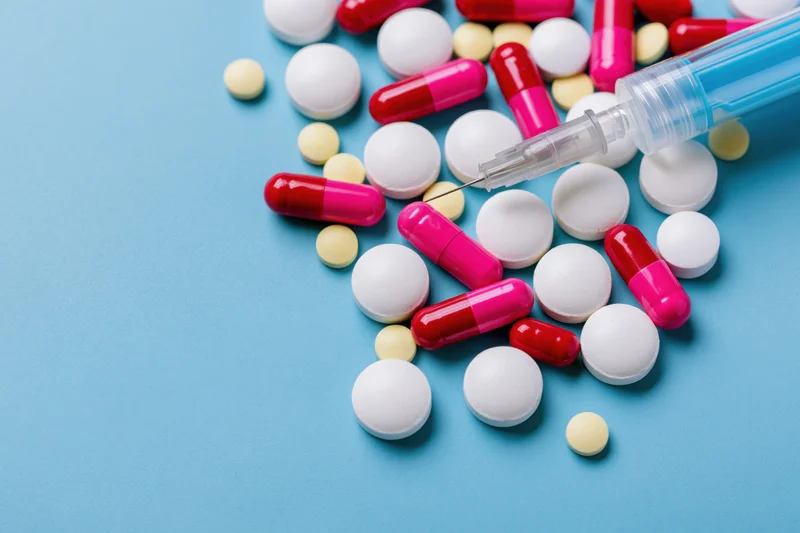 Assorted pharmaceutical pills and capsules scattered beside a syringe on a blue surface
