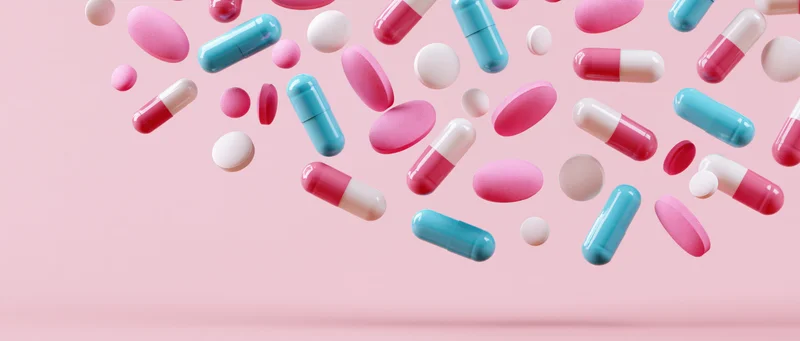 Assorted pharmaceutical pills and capsules floating mid-air against a pink background
