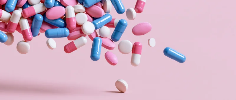 Assorted pharmaceutical pills and capsules scattered mid-air against a pink background