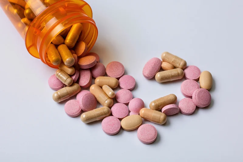 Assorted pills and capsules arranged in heart shape on white surface from orange bottle