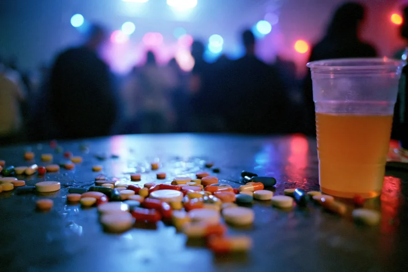 Scattered colorful pills and capsules on a table beside a plastic cup of drink at a party with blurred people and neon lights in the background