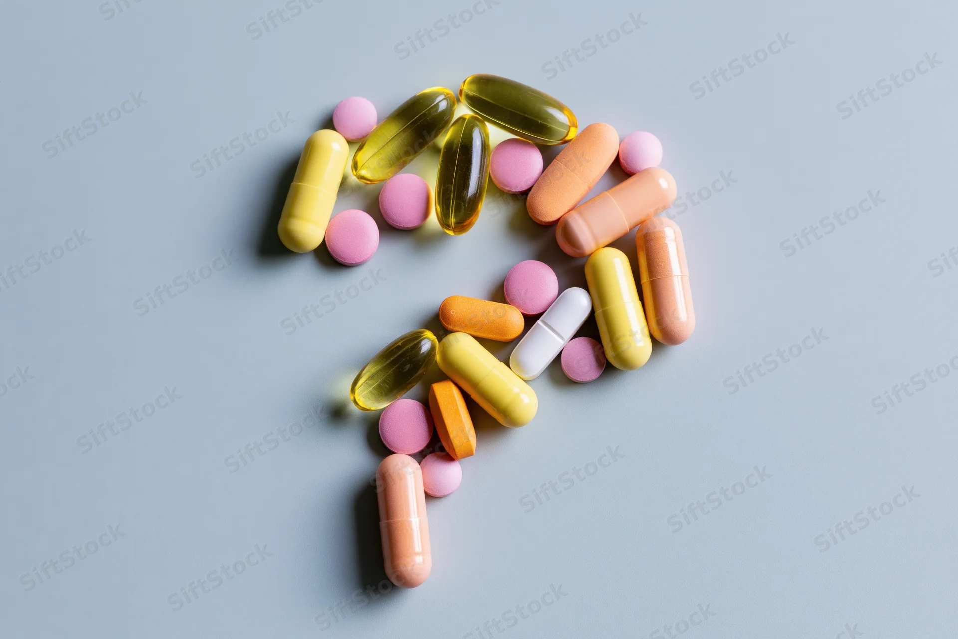 Assorted pills and capsules arranged in the shape of a question mark on a blue surface