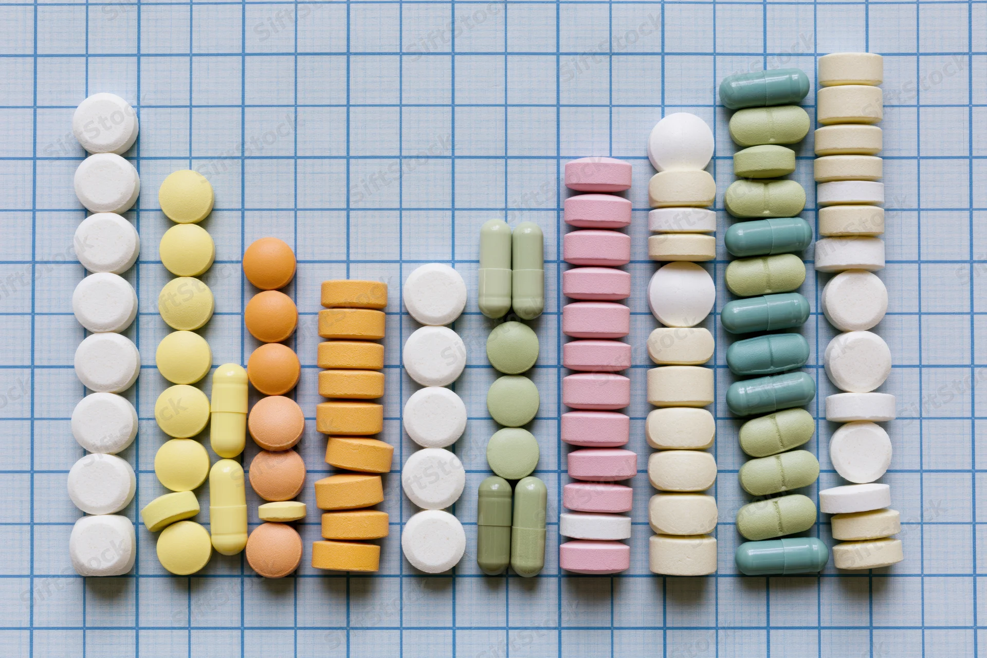 Assorted prescription pills and capsules arranged in a rising bar graph pattern on a blue grid background