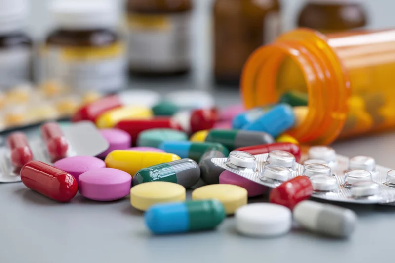 Assorted prescription pills and capsules scattered across a surface with an orange bottle and blister packs nearby