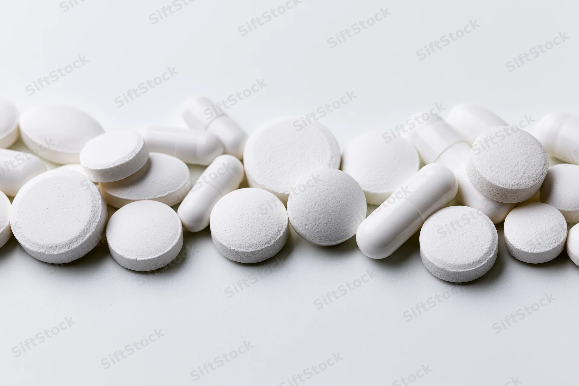 Assorted white pharmaceutical pills and capsules scattered across a plain white surface