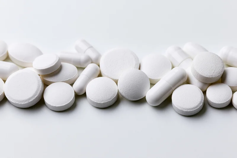 Assorted white pharmaceutical pills and capsules scattered across a plain white surface