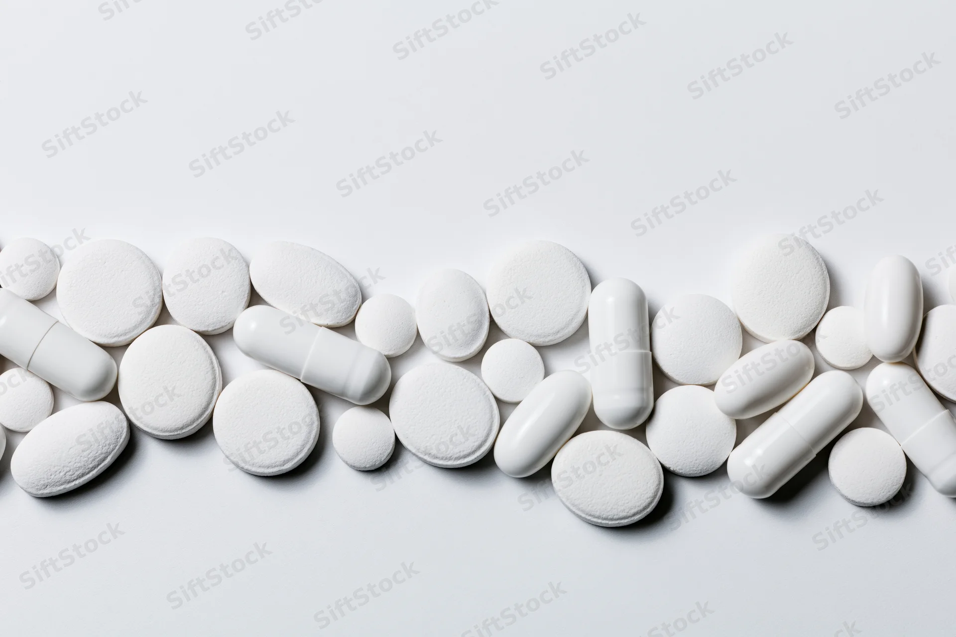 Assorted white pharmaceutical pills and capsules scattered across a plain white surface