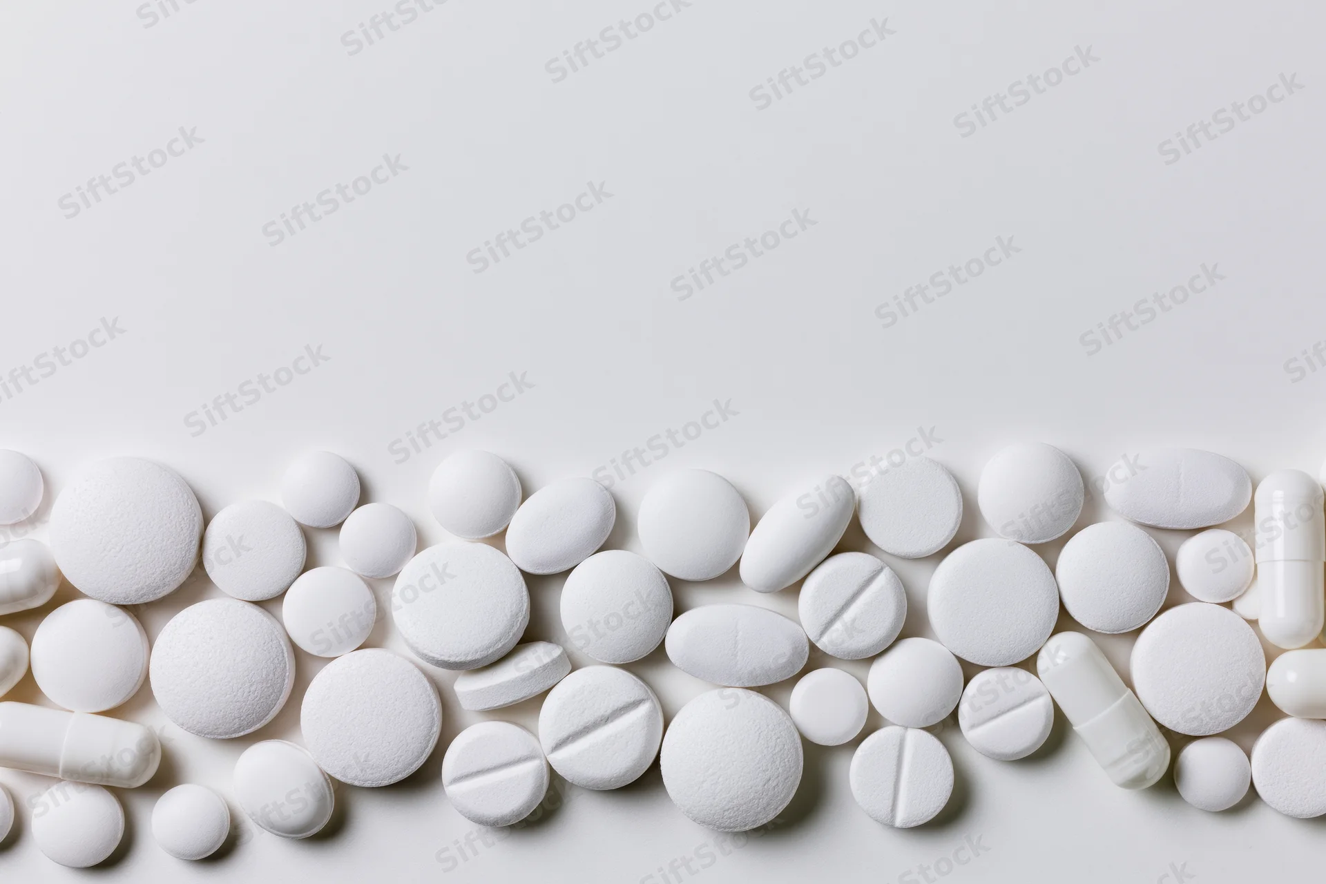 Assorted white pharmaceutical tablets and capsules scattered along the bottom of a plain white surface