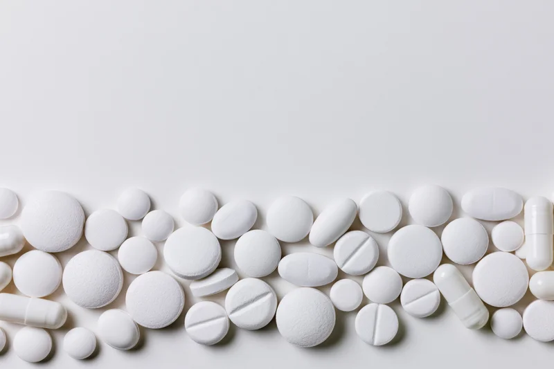 Assorted white pharmaceutical tablets and capsules scattered along the bottom of a plain white surface