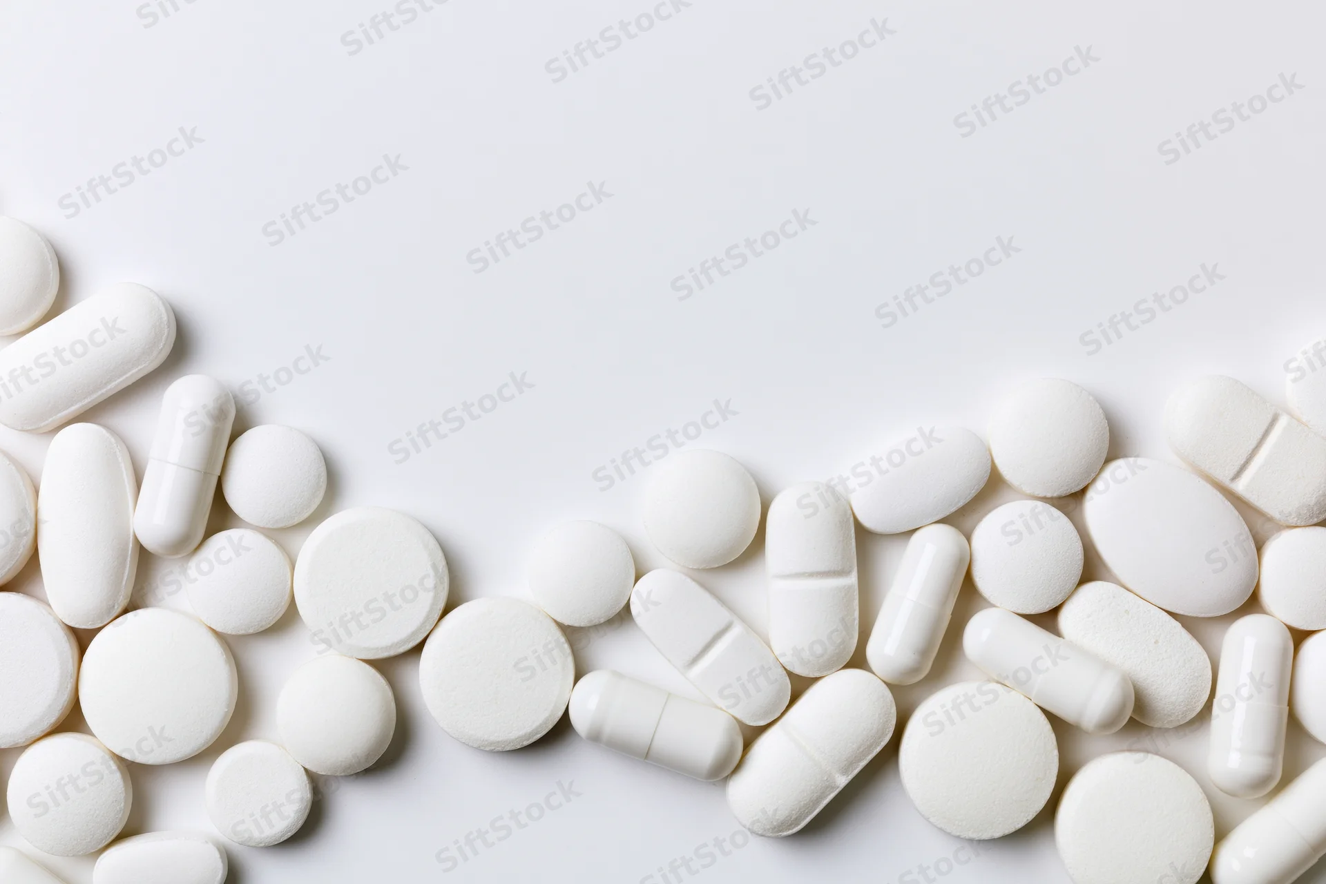 Assorted white pharmaceutical tablets and capsules scattered across a plain white surface with copyspace