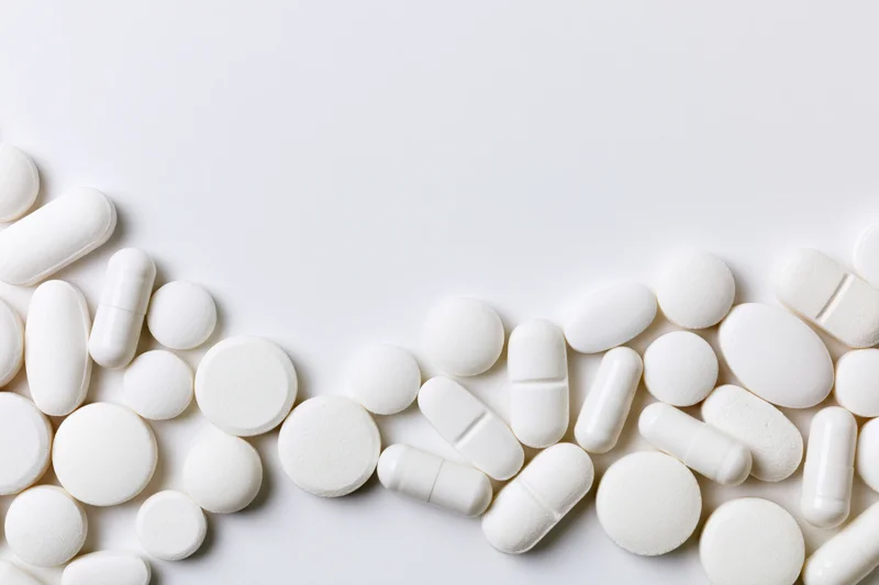 Assorted white pharmaceutical tablets and capsules scattered across a plain white surface with copyspace