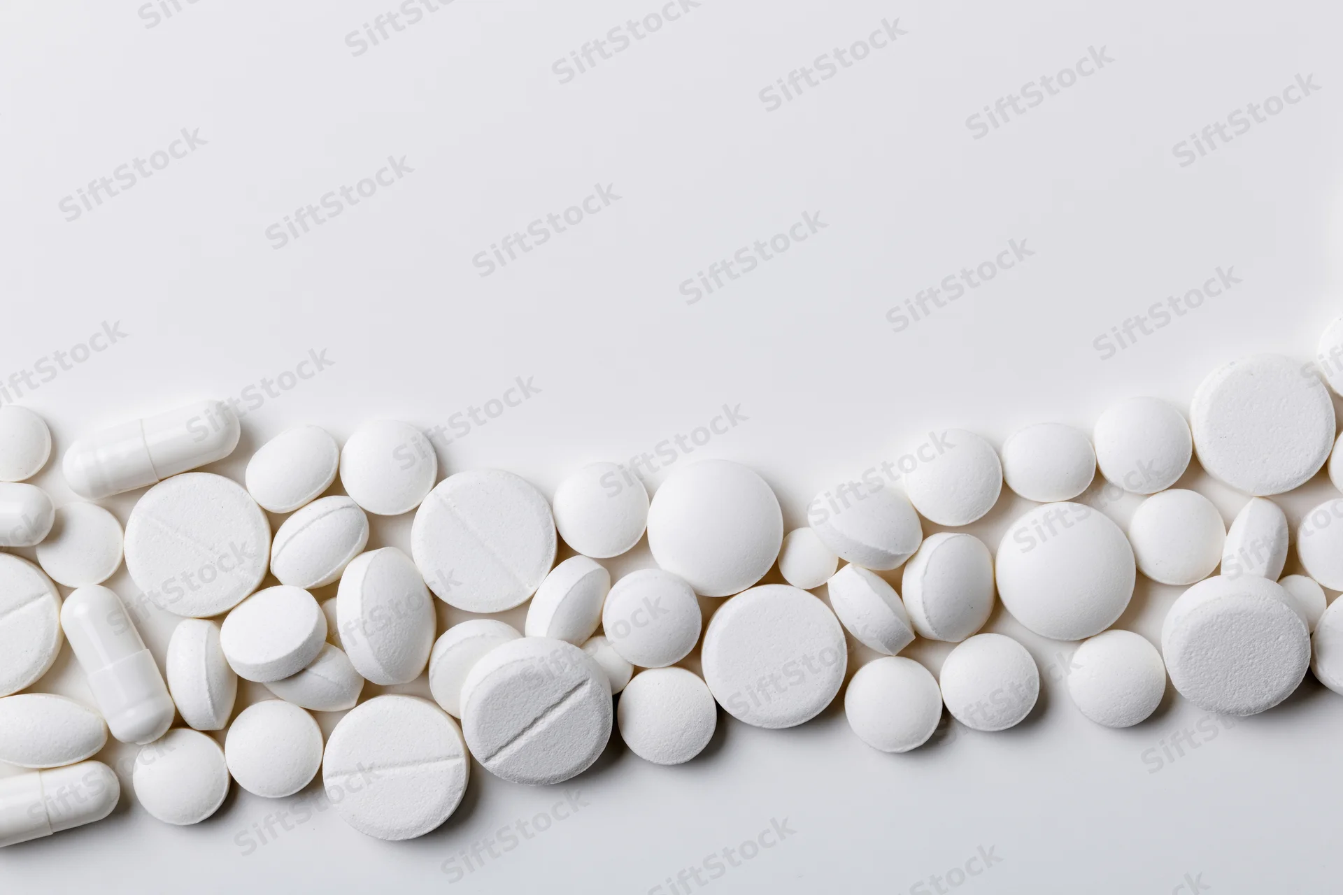Assorted white pharmaceutical tablets and capsules scattered across a plain white surface