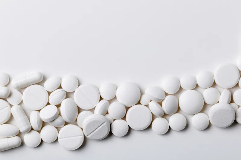 Assorted white pharmaceutical tablets and capsules scattered across a plain white surface