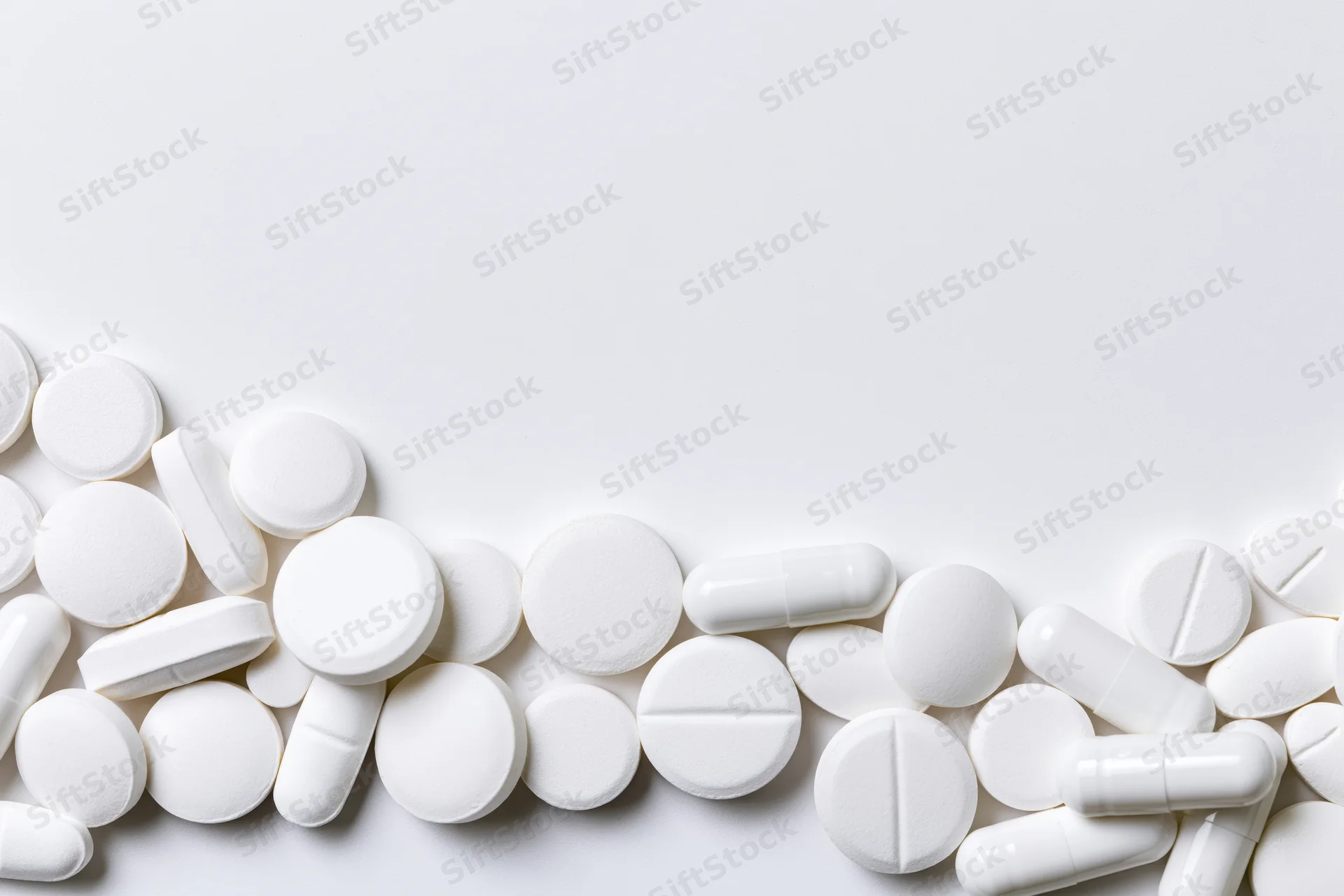 Assorted white pharmaceutical tablets and capsules scattered along the bottom of a plain white surface