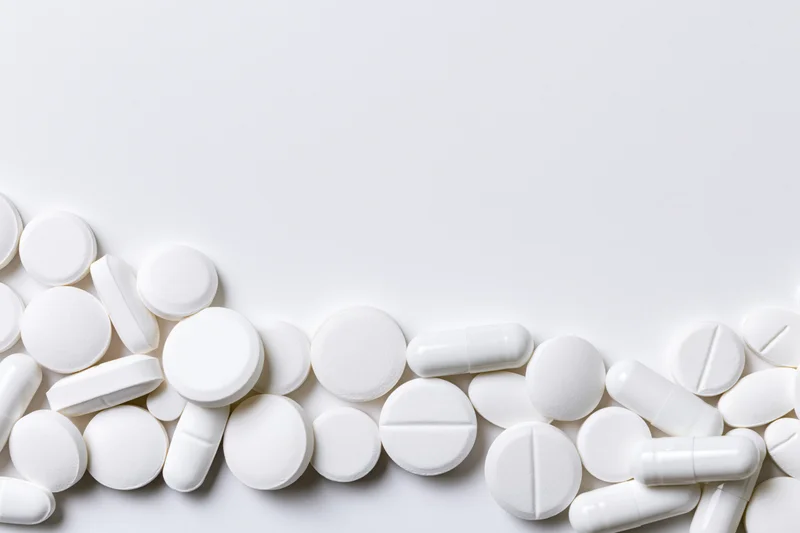 Assorted white pharmaceutical tablets and capsules scattered along the bottom of a plain white surface
