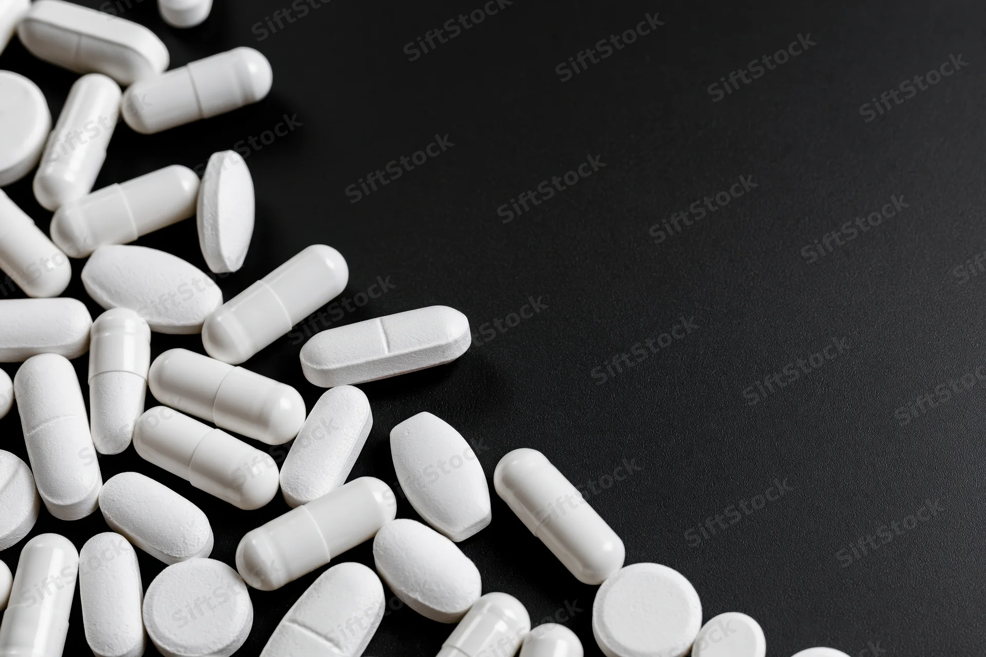 White pills and capsules scattered loosely on black surface with copyspace