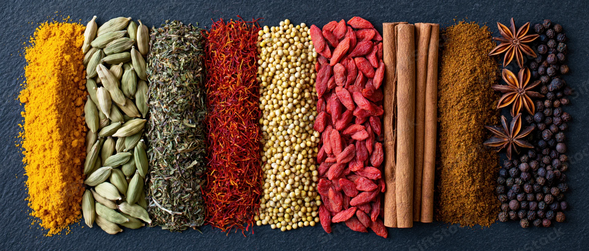 Assorted spices arranged in parallel rows on dark slate surface