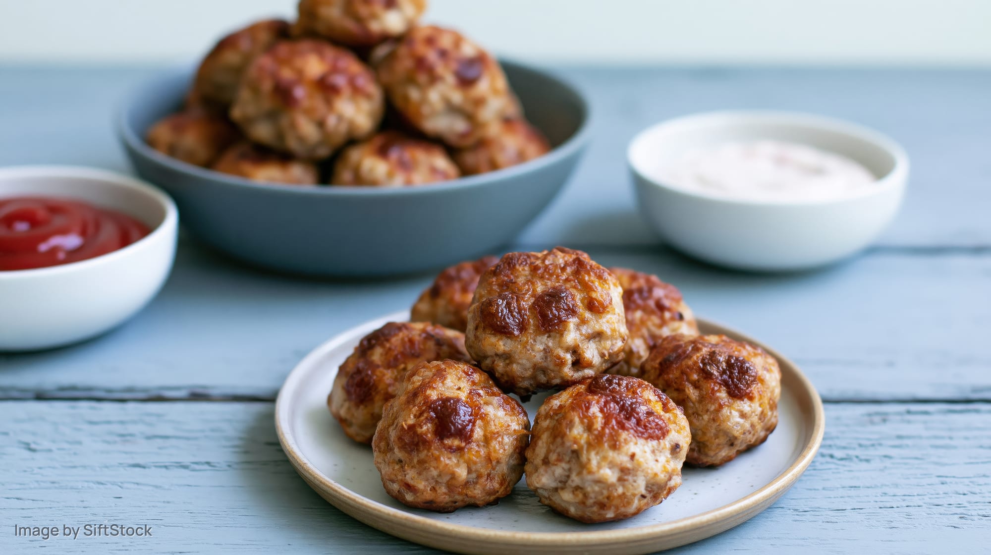 Baked meatballs arranged on a small ceramic plate with ketchup and creamy dip on a blue wooden table