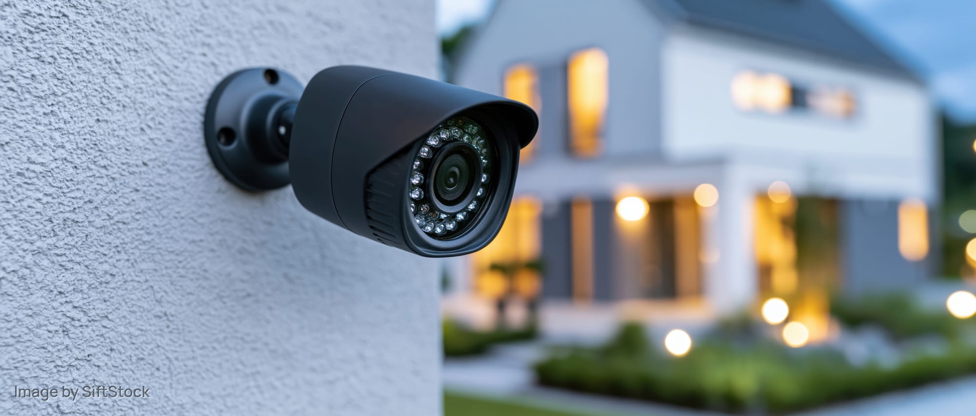 Black bullet-style security camera mounted on a textured wall outside a modern house at dusk