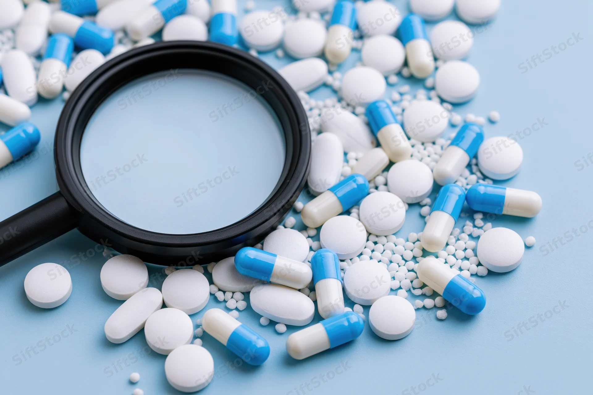 Magnifying glass resting on scattered white and blue pills on a light blue surface