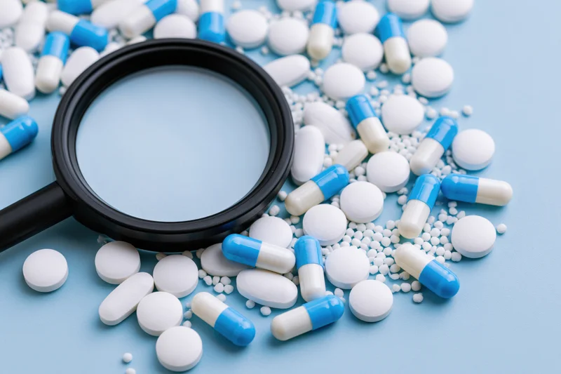 Magnifying glass resting on scattered white and blue pills on a light blue surface