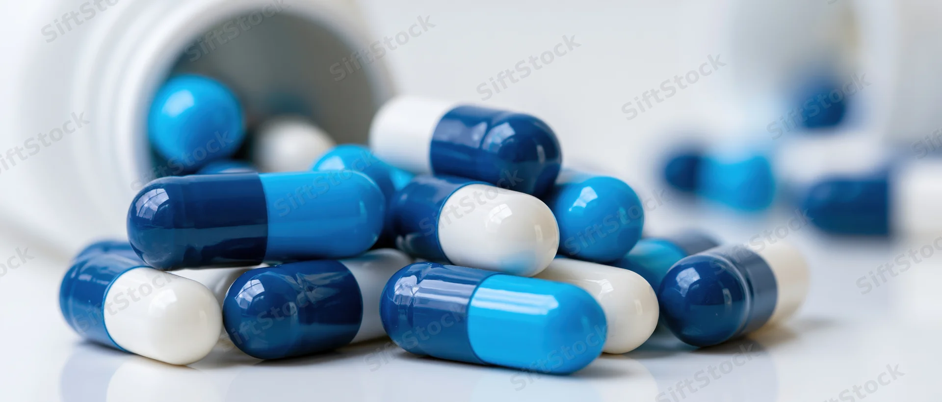 Blue and white medication capsules scattered on a white surface near a bottle opening