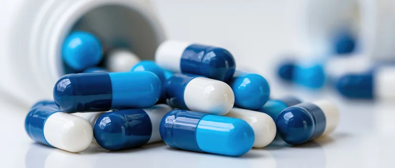 Blue and white medication capsules scattered on a white surface near a bottle opening