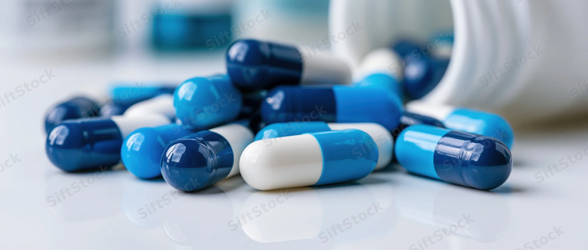 Blue and white medication capsules scattered on a reflective white surface near a white bottle