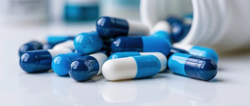 Blue and white medication capsules scattered on a reflective white surface near a white bottle