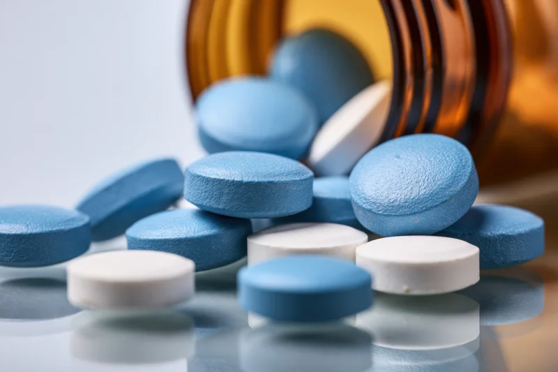 Blue and white round pharmaceutical tablets spilled from an amber glass bottle onto a reflective surface