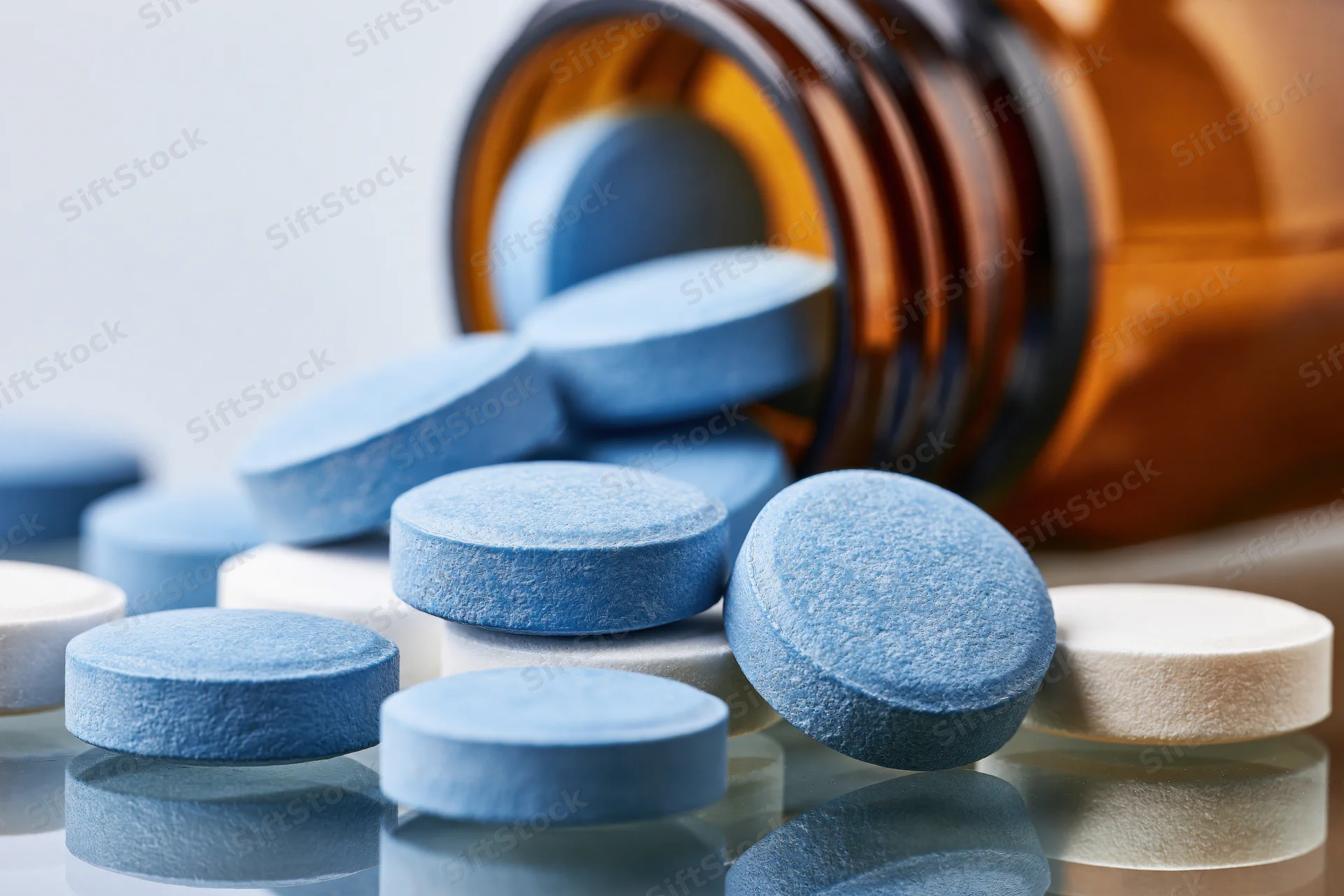 Blue and white round tablets spilled from an amber glass bottle on a reflective surface