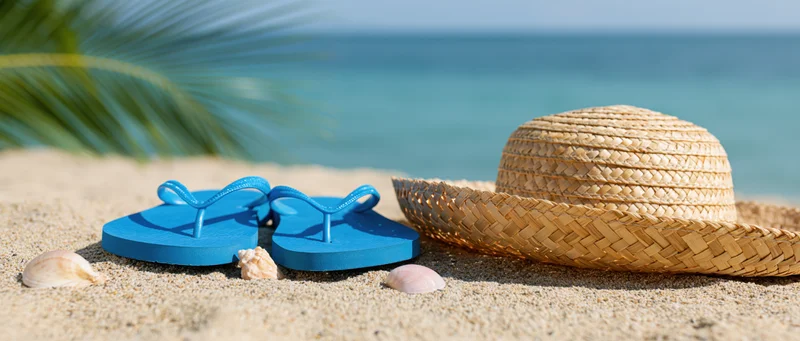 Blue flip-flops and woven straw hat with seashells on sandy beach near ocean