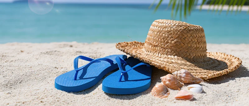 Blue flip flops and a woven straw hat with seashells on sandy beach near ocean