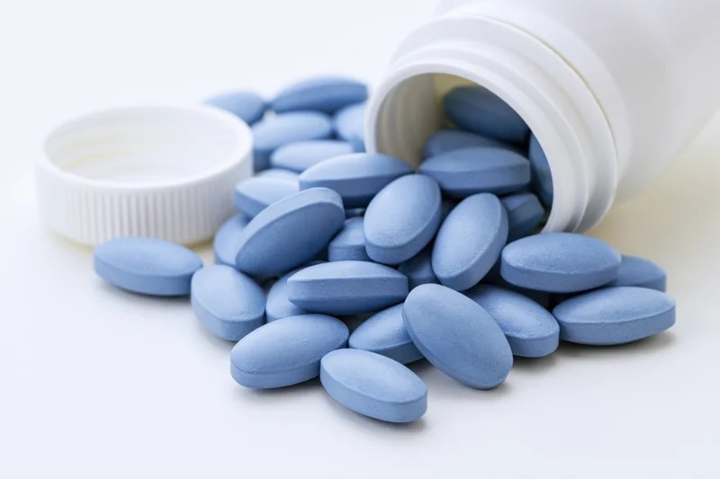 Blue oval pills spilling from a tipped white plastic bottle on a white surface