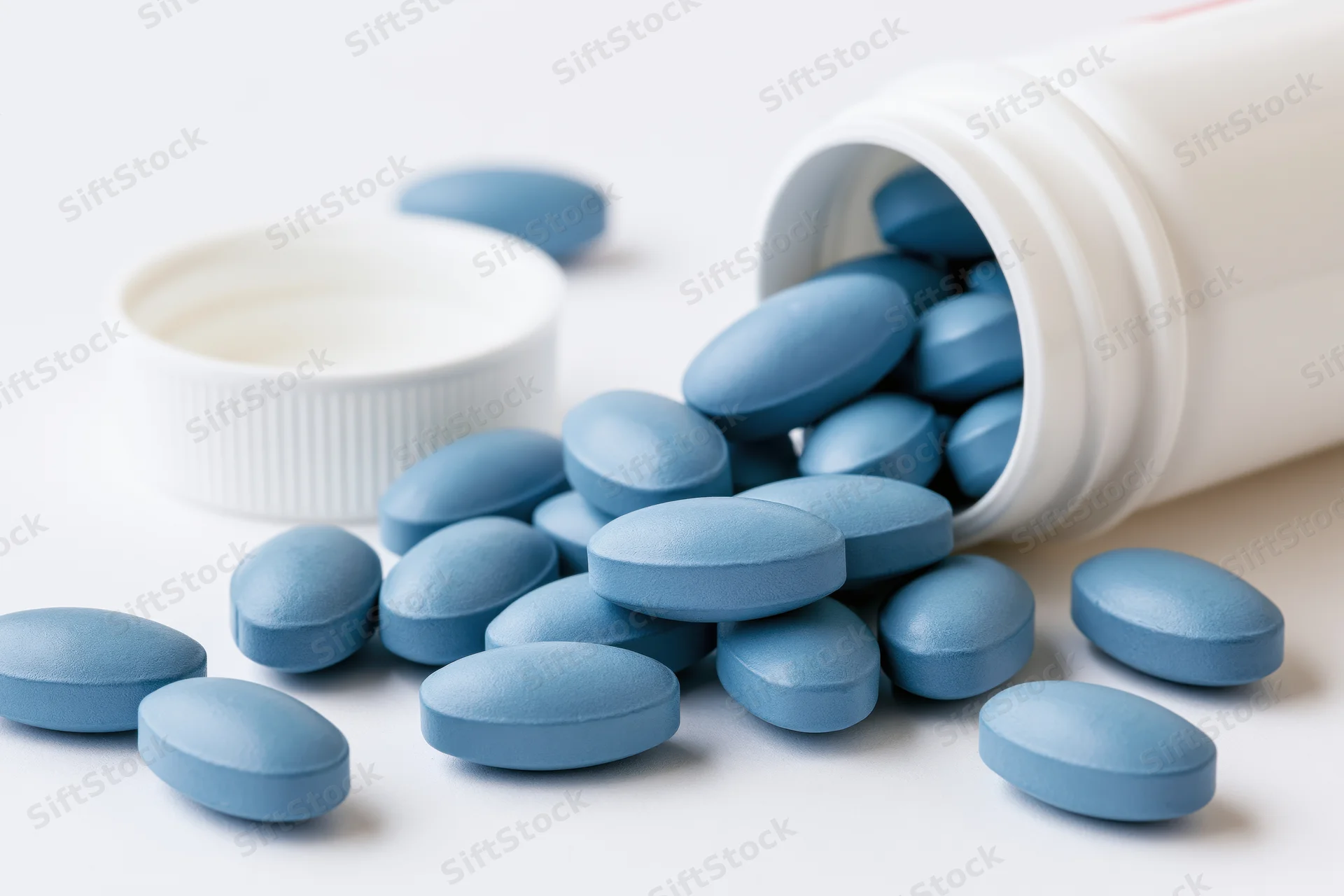 Blue oval pills spilling from a tipped white plastic bottle on a plain surface
