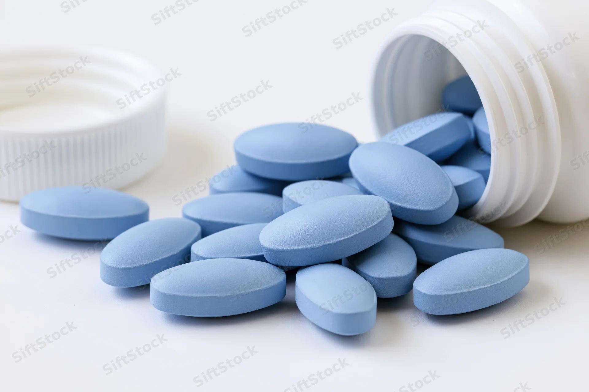 Blue oval pills spilling from a tipped white plastic bottle on a white surface