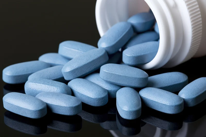 Blue oval pills spilling from a white plastic bottle onto a reflective surface