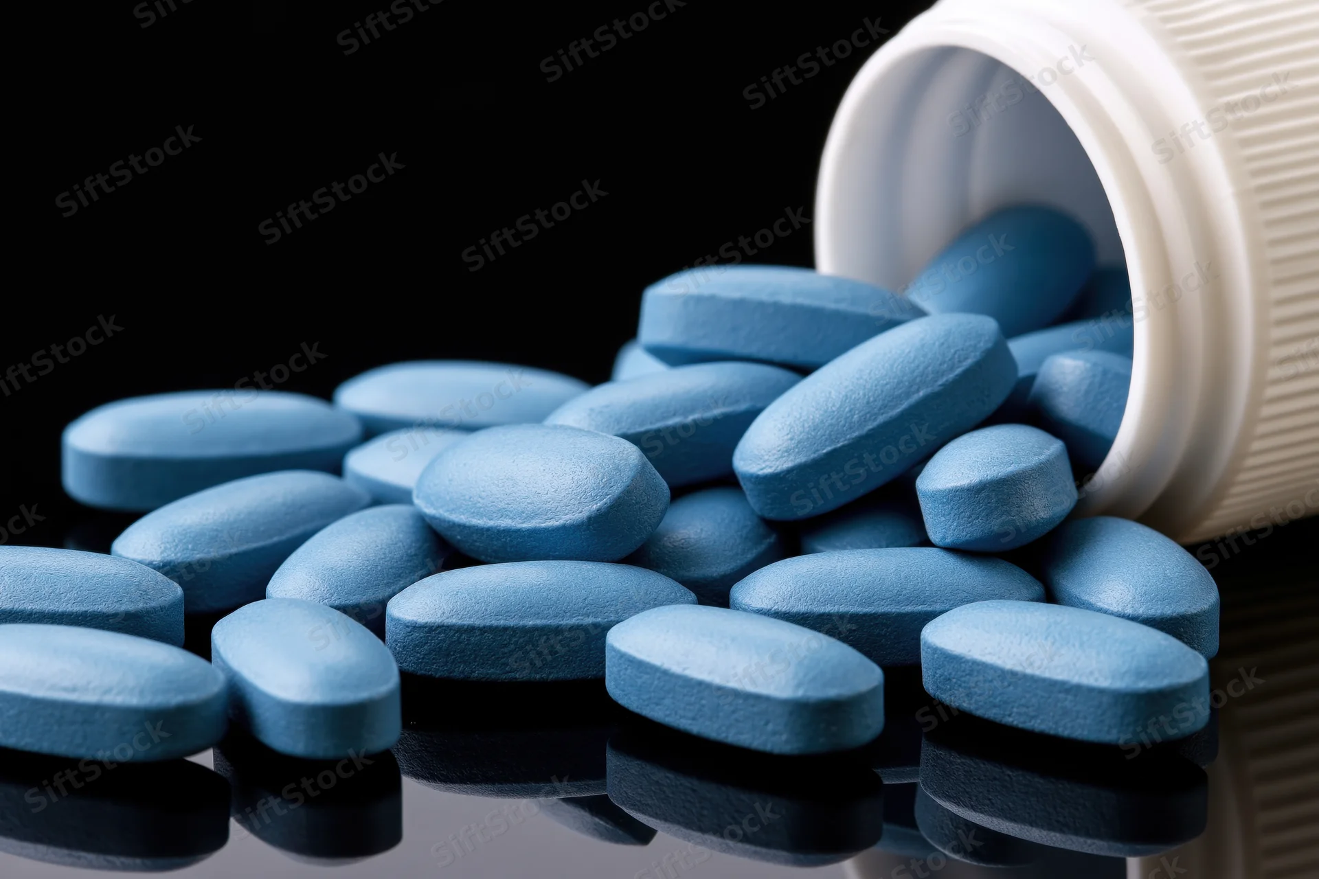 Blue oval pills spilling from a tipped white plastic bottle onto a reflective black surface