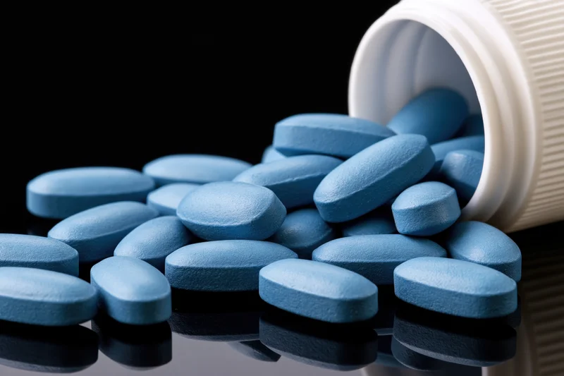 Blue oval pills spilling from a tipped white plastic bottle onto a reflective black surface