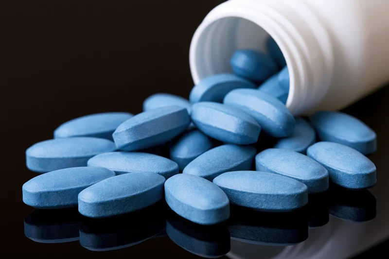 Blue oval pills spilling from a white plastic bottle onto a dark reflective surface