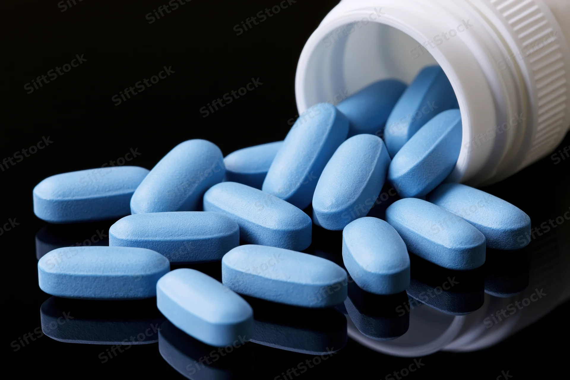 Blue oval pills spilling from a white plastic bottle onto a reflective surface